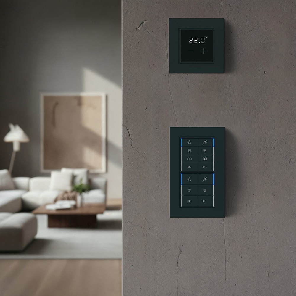 Schwarzer KNX kallysto.pro Taster Schalterkombination und schwarzer Raumthermostat von Hager auf grauer Betonwand mit Wohnzimmer im Hintergrund