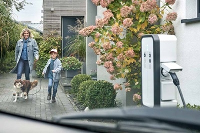 Elektrofahrzeug-Ladestation an einer privaten Einfahrt mit einer Person und einem Kind, die im Hintergrund einen Hund ausführen