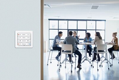 Gruppe von Mitarbeitern in einem Meeting-Raum. Im Vordergrund ein Berker Tastsensor 4fach Komfort, Q.1 polarweiß, mit individuell lackierten Schriftfeldern und gelaserter Beschriftung.