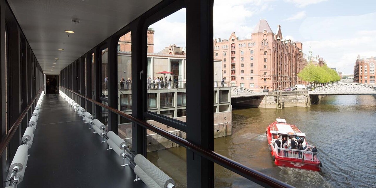 Blick von einem überdachten Gehweg mit Aussicht auf einen Kanal, eine Brücke und traditionelle Gebäude, vorbeifahrendes Ausflugsboot