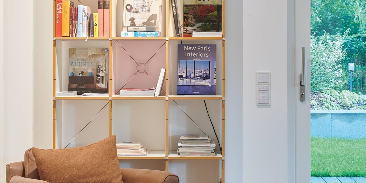 Modernes Zuhause mit Bücherregal, komfortablem Sofa und an der Wand montiertem Hager Hausautomation-Bedienfeld neben der Tür