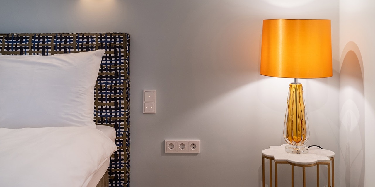 Modernes Schlafzimmer mit weißer Bettwäsche, dekorativem Kopfteil, Wandlichtschaltern, Steckdosen und einer orangen Lampe auf einem Nachttisch