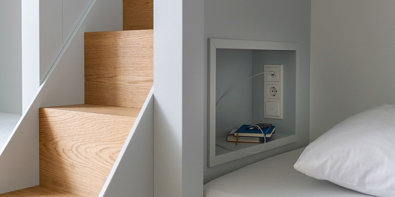 Modernes Interieur mit Holztreppe und eingebauter Steckdose an der Wand neben dem Bett mit aufladendem Handy