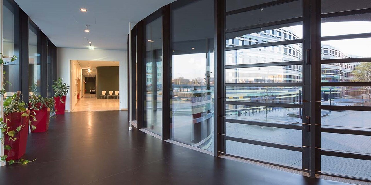 Moderner Bürorflur mit Glaswänden, Zimmerpflanzen und an der Decke montierten Leuchten, die sich auf dem Boden spiegeln