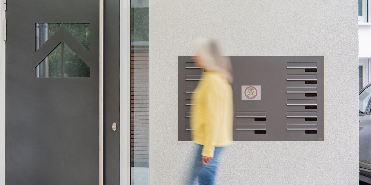 Moderner Hauseingang mit unscharfer Person, die an einem Klingeltableau und Briefkastenschlitzen vorbeigeht