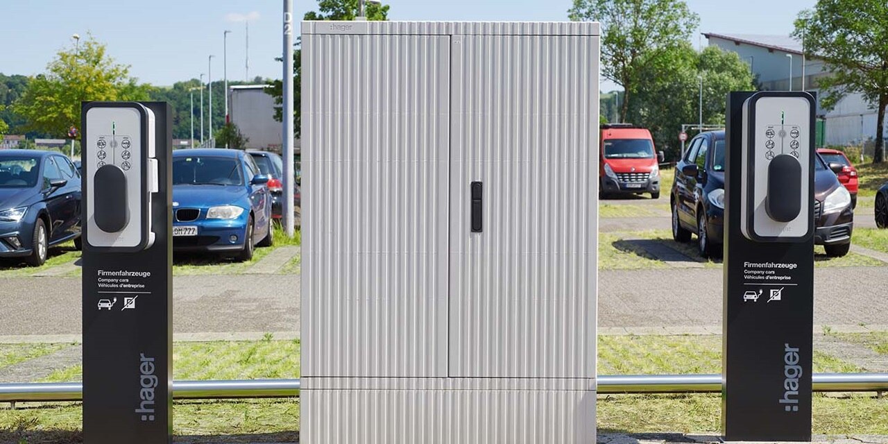 Hager-markierte Ladestationen für Elektrofahrzeuge auf einem Parkplatz mit Autos und Grünanlagen im Hintergrund