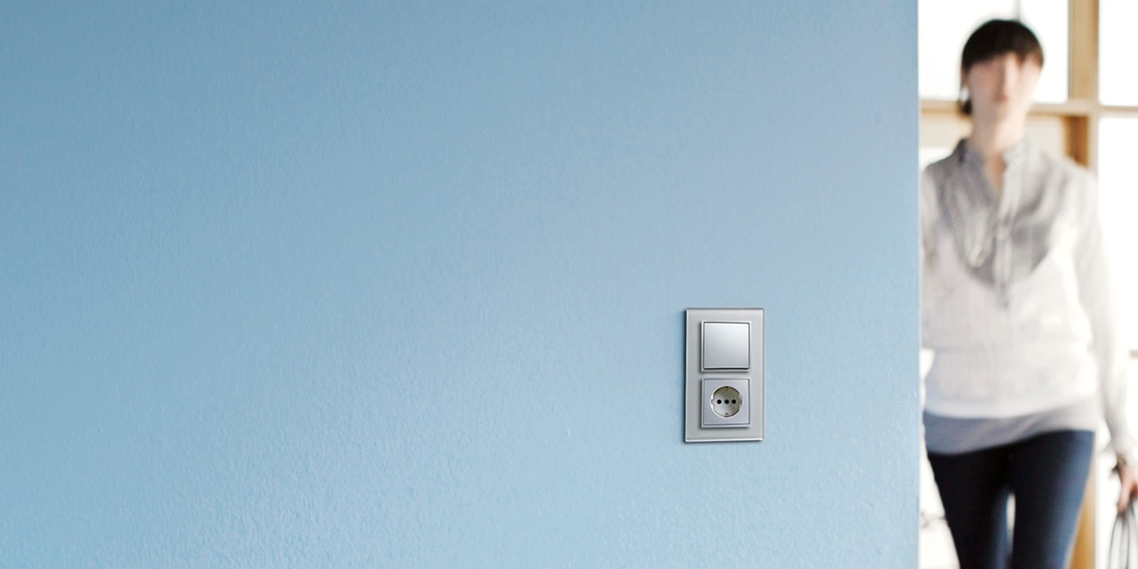 Moderner Lichtschalter und Steckdose an einer blau gestrichenen Wand mit einer unscharfen Person im Hintergrund.