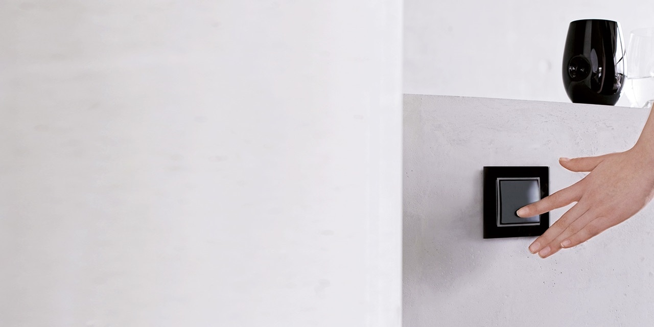 Person streckt sich nach einem schwarzen Lichtschalter an einer weißen Wand, minimalistisches Design