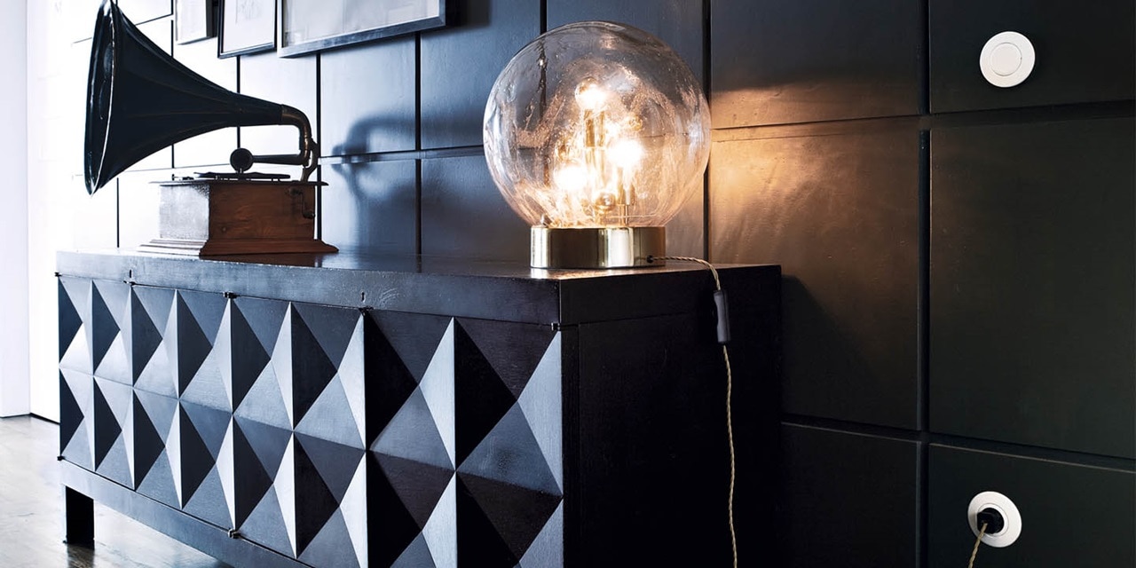 Vintage-Glühbirne mit sichtbaren Glühfäden auf Messingsockel, neben einem Grammophon auf einem Schrank mit geometrischem Muster, mit einer runden Wand-Lichtschalter