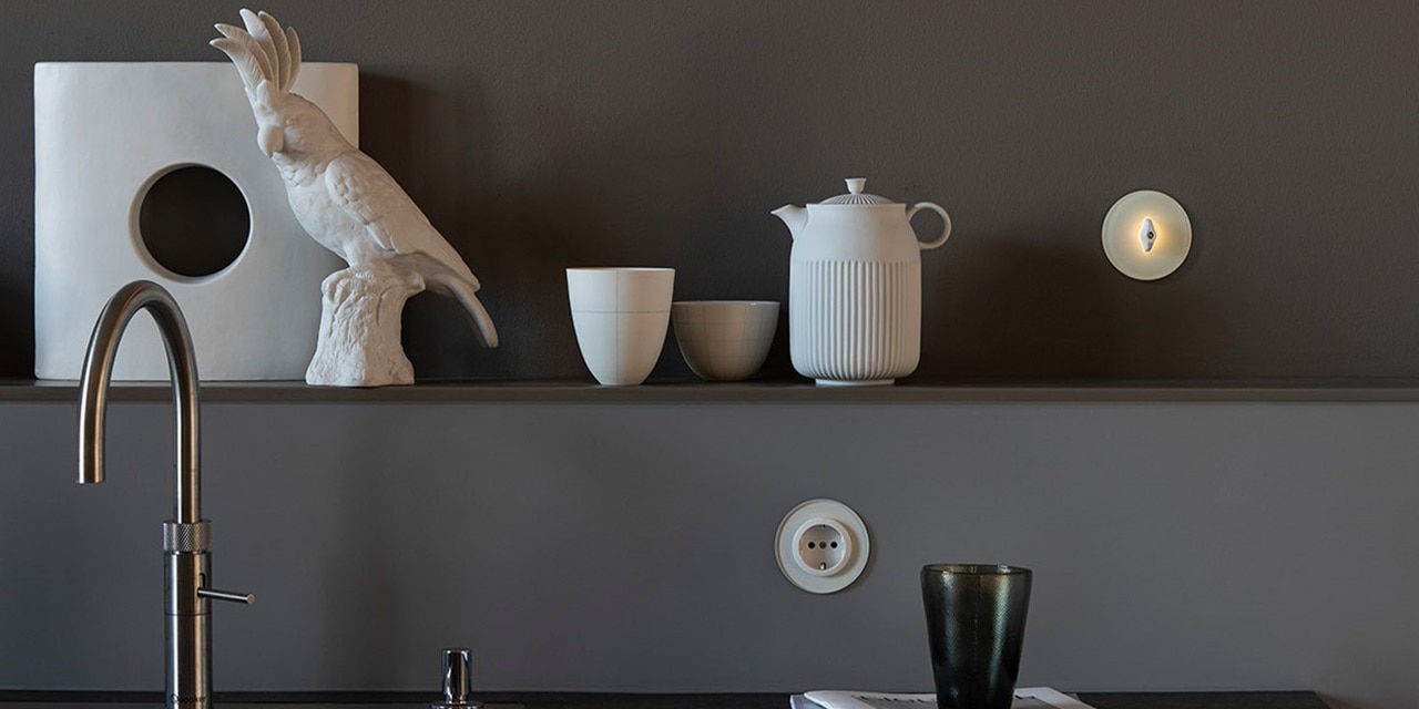 Stilvolles Kücheninterieur mit einer Keramikvogelskulptur, Teekanne, Tassen und einem modernen Lichtschalter an der Wand