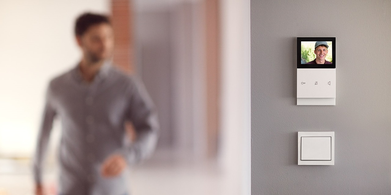 Videogegensprechanlage mit Bildschirm an einer Wand in einem modernen Wohnraum, unscharfe Person im Hintergrund