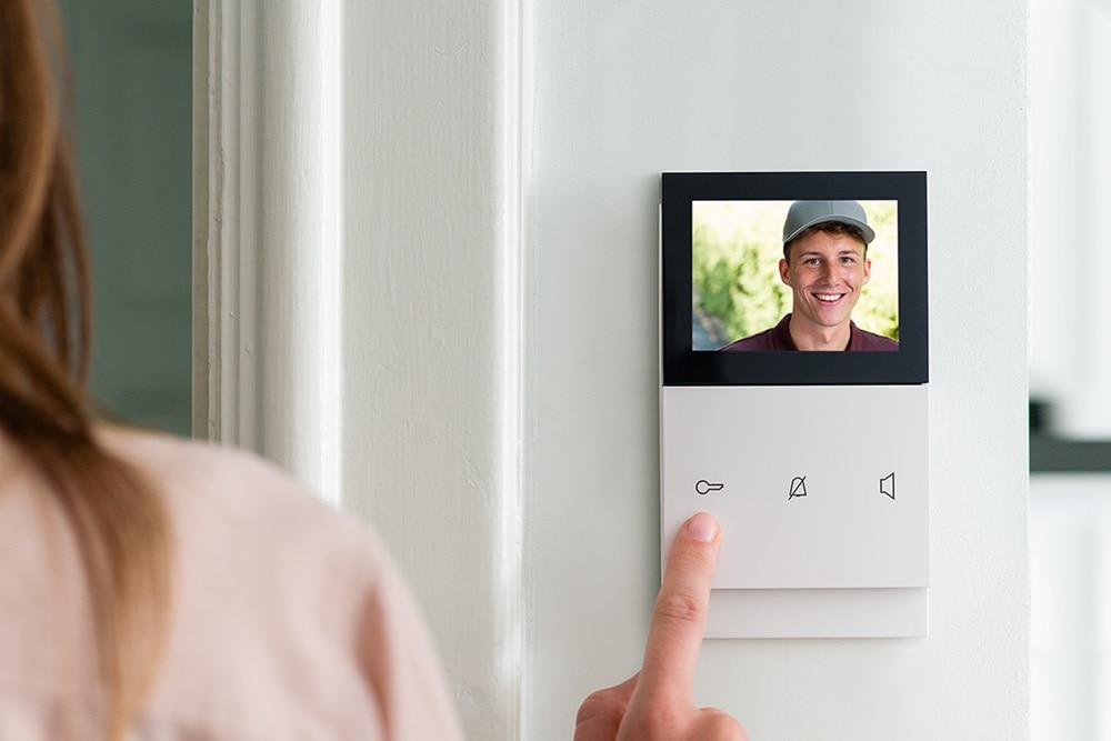 Weiße Video-Innenstation von Elcom mit Bild einer Person vor der Tür auf dem Display, Montage Aufputz auf weißer Wand
