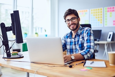 Lächelnder Mann mit Vollbart am Schreibtisch sitzend, vor ihm ein aufgeklapptes weißes Notebook