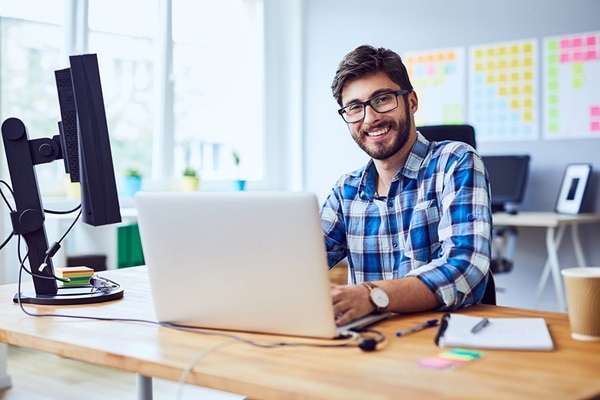 Lächelnder Mann mit Vollbart am Schreibtisch sitzend, vor ihm ein aufgeklapptes weißes Notebook