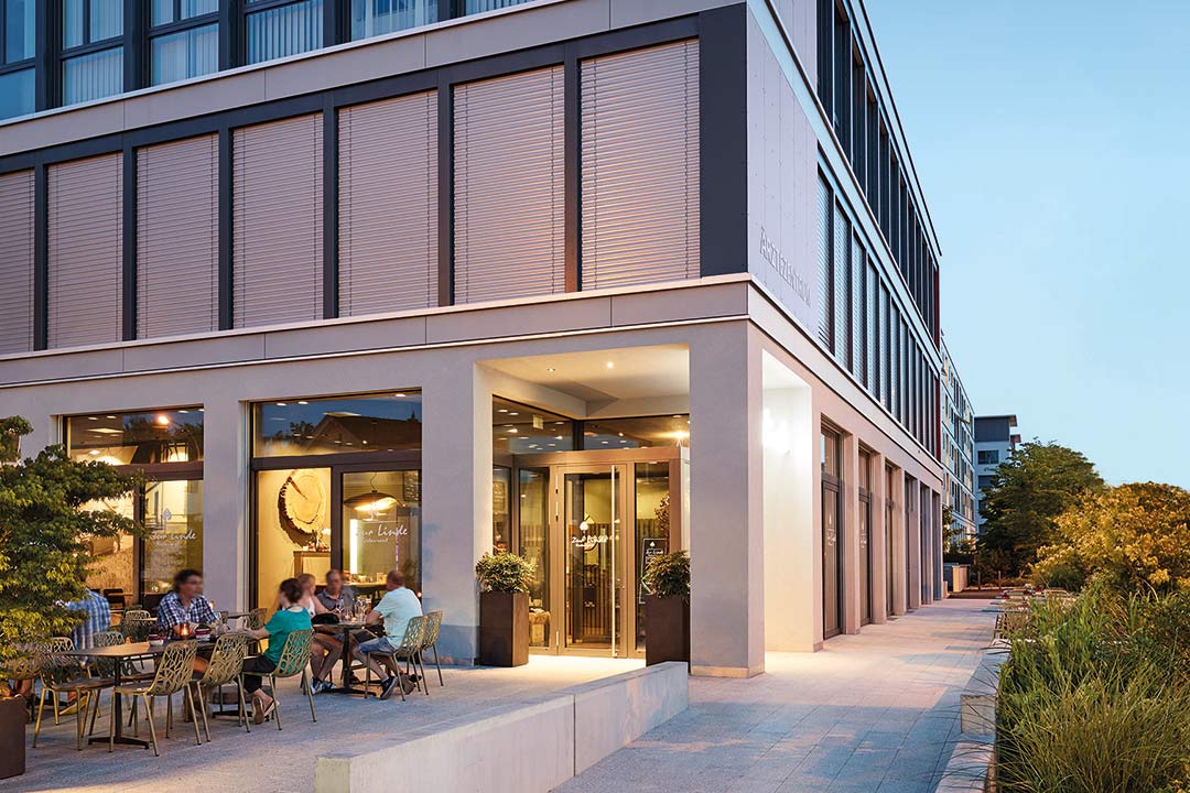 Blick auf modernes Gebäude am Abend mit beleuchtetem Restaurant im EG und Gästen an Tischen im Freien