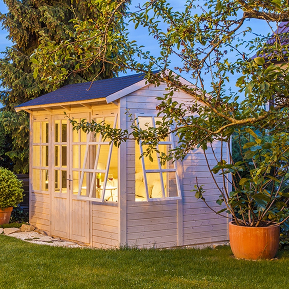 Blick auf Gartenhaus aus hellem Holz mit aktivierter Innenbeleuchtung umgeben von Gras und Sträuchern