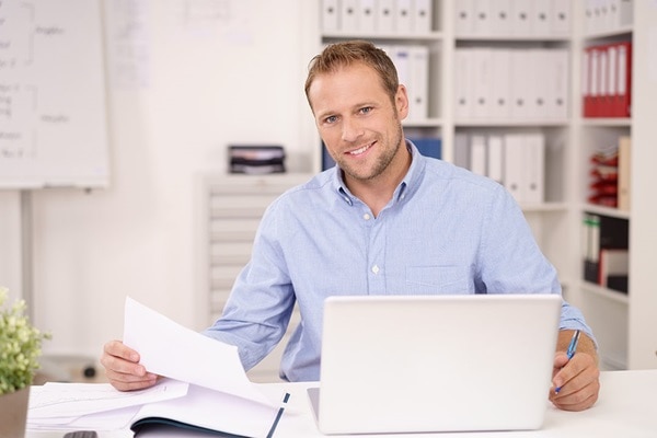 Mann in Büro vor Notebook sitzend mit Blatt Papier in der Hand