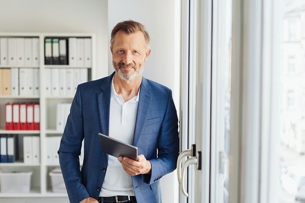 Mann in Büro stehend mit Tablet in der Hand, Körper gegen Terrassentür gelehnt
