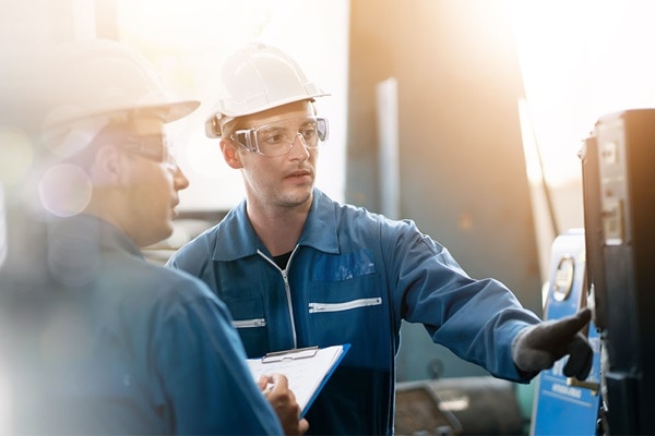 Zwei Männer in Schutzkleidung vor Maschinen, einer von ihnen führt Bedienung durch