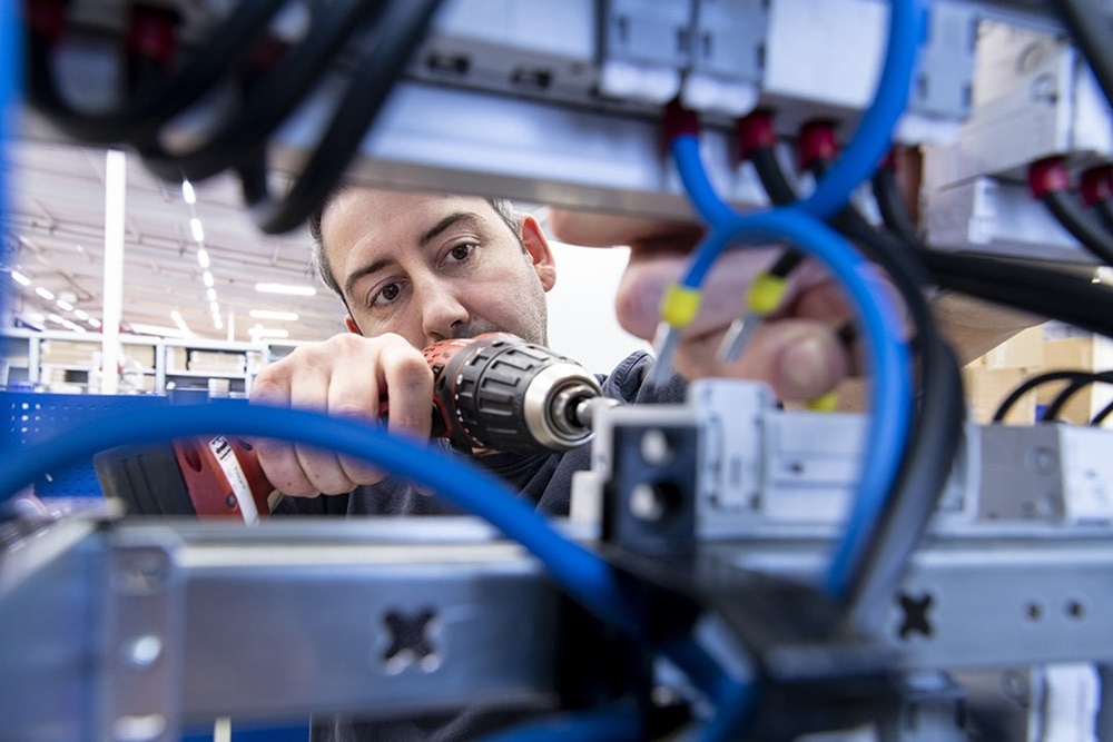 Blick durch offenen Wandlerschrank hindurch auf dahinter stehenden Elektrikter, der mit Akkuschrauber Schraubklemmen befestigt