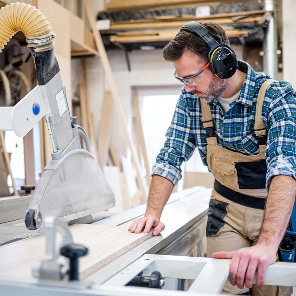 Schreiner in Werkstatt/Werkraum schneidet mit konzentriertem Blick Holzbrett mit Tischkreissäge