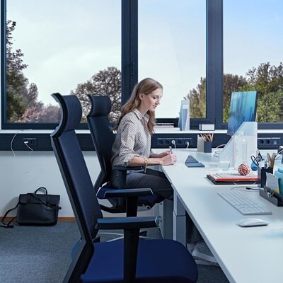 Blick in Büro mit Frau vor Schreibtisch, im Hintergrund ist ein Hager tehalit Brüstungskanal in Blau zu sehen