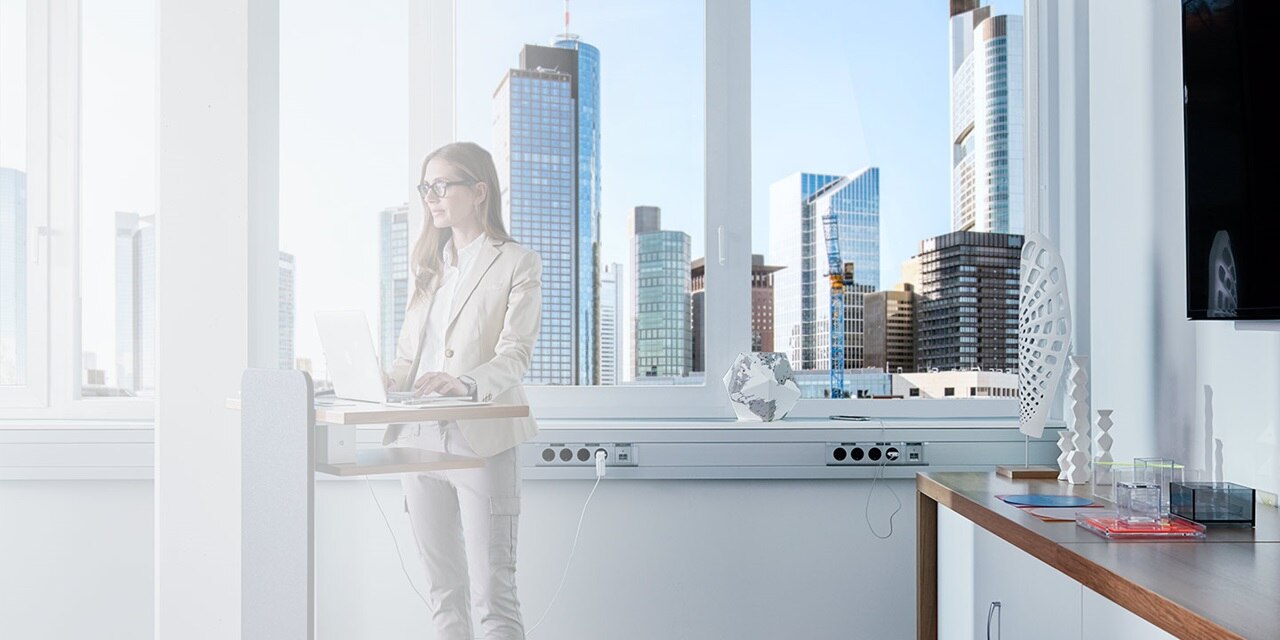 Person benutzt einen Stehschreibtisch mit Laptop in einem modernen Büro mit Stadtblick und einer Steckdosenleiste unter dem Tisch