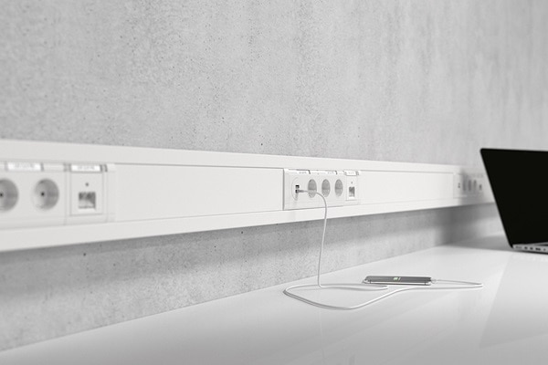 Schrägansicht Hager Brüstungskanal an Sichtbetonwand, u.a. mit USB-Dose und eingestecktem Ladekabel für Smartphone