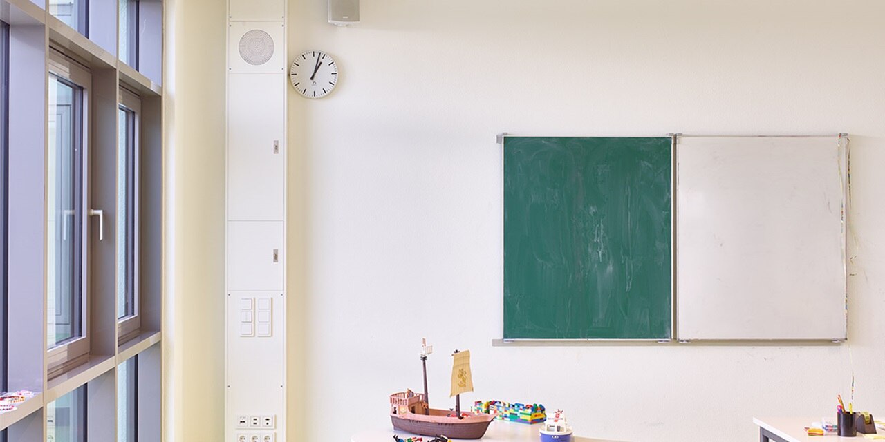 Blick in Klassenraum einer Grundschule mit Hager Mediensäule in Weiß links, rechts daneben Tafeln und Tische mit Spielzeug und Malstiften 