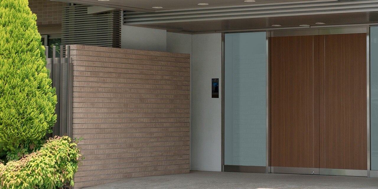 Blick auf überdachten Hauseingang mit Hager intercom motion Video-Außenstation in schwarz links neben der holzfarbenen Hauseingangstür