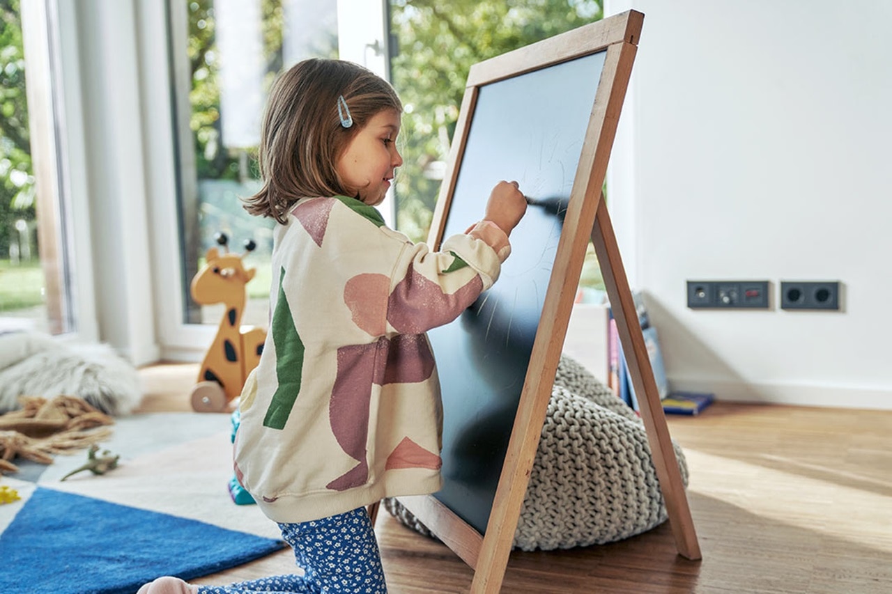 Kind malt auf einer Tafel mit Hager Steckdosen und Schaltern im Hintergrund