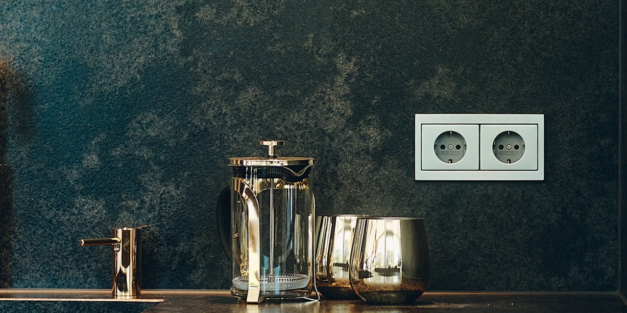 Modernes Kücheninterieur mit einer French Press, Gläsern und einer stilvollen Doppelsteckdose an einer dunkelblauen Wand