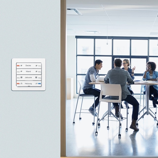 Intelligentes Steuerungspanel für das Smart Home an einer Wand in modernem Büroumfeld mit Personen im Hintergrund bei einem Meeting