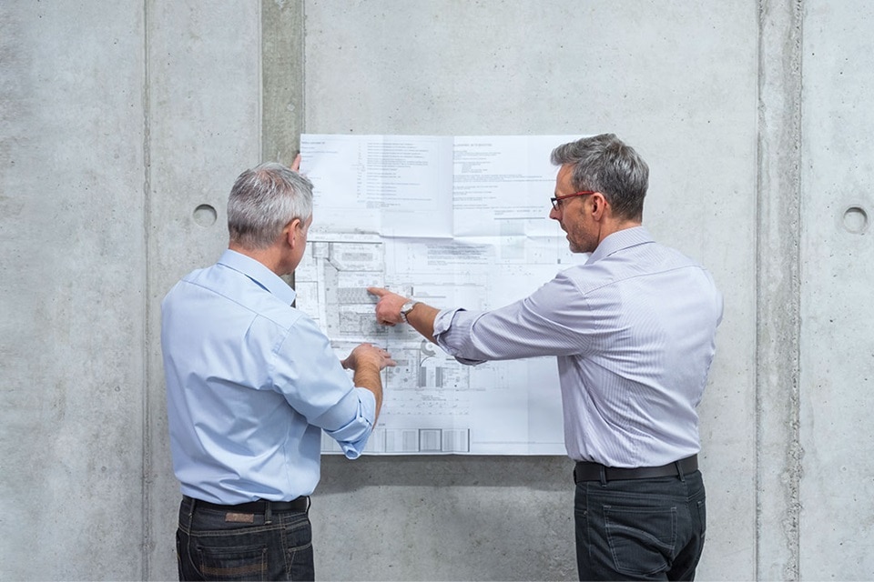 Zwei Fachleute diskutieren über einen Bauplan an einer Betonwand, konzentriert auf die Details des Elektroplans