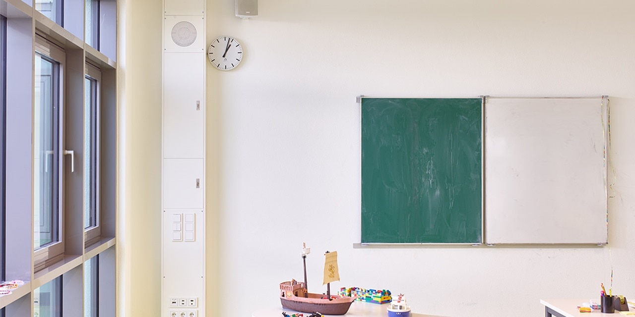Klassenzimmer-Innenansicht mit Tafel und Whiteboard nebeneinander an der Wand, Uhr darüber und Elektroverteilungstafel links
