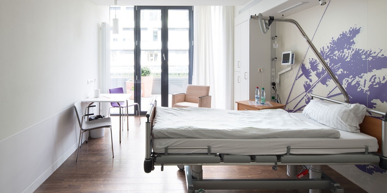 Modernes Krankenzimmer mit verstellbarem Bett, Nachttisch, Stühlen und großen Fenstern, die zu einem Balkon führen