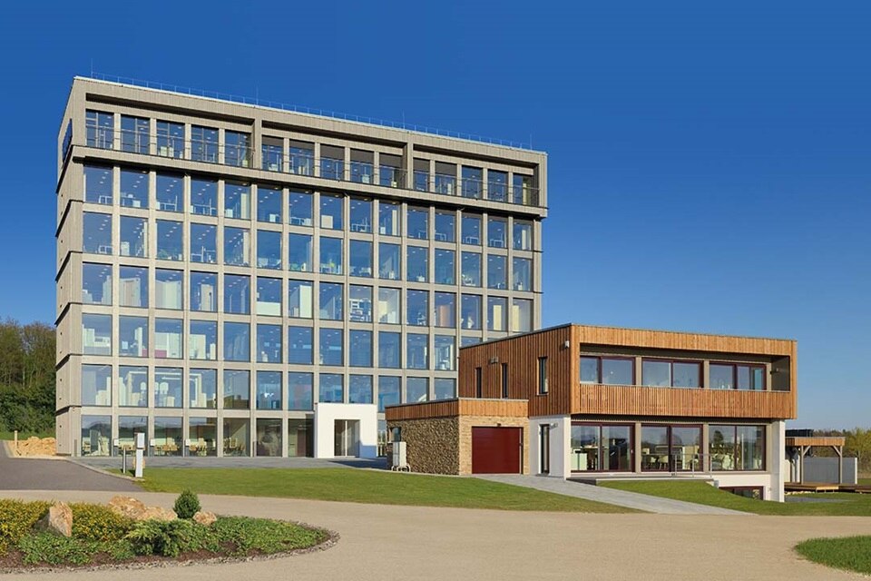 Modernes Geschäftsgebäude mit Glasfassade und angrenzendem Holzbau unter klarem blauem Himmel