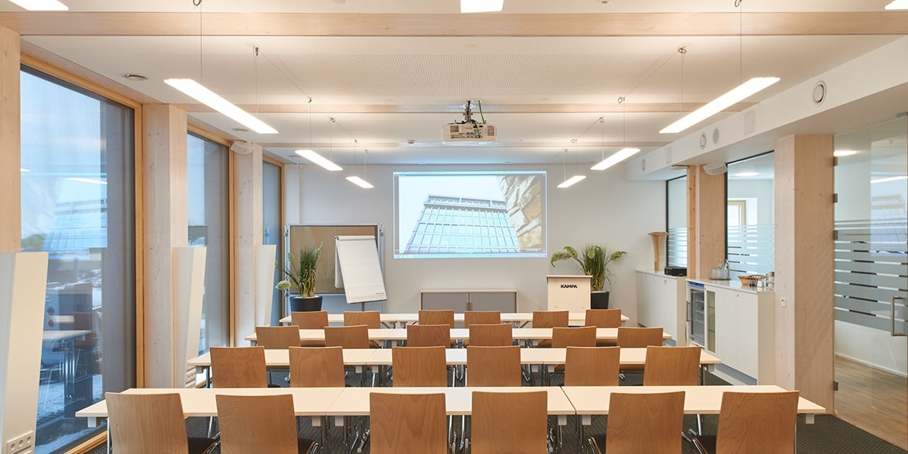 Moderner Konferenzraum mit Reihen von Holzstühlen, einer Projektionsleinwand und eingebauten Beleuchtungskörpern