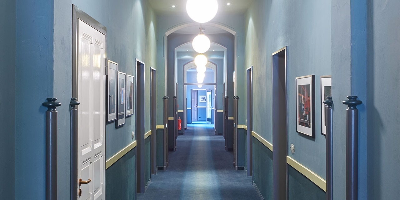 Innenansicht eines Korridors mit blauen Wänden, hängenden Bildern, weißen Türen und Deckenleuchten