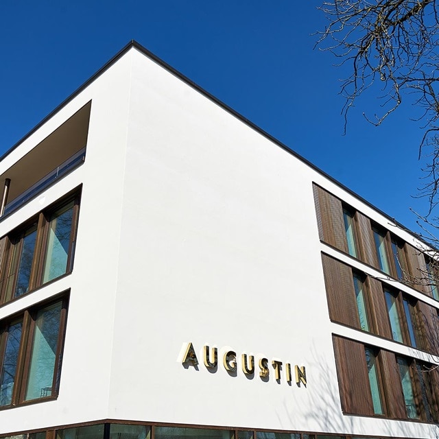 Moderne Gebäudefassade mit 'AUGUSTIN'-Beschriftung und großen Fenstern vor klarem blauem Himmel