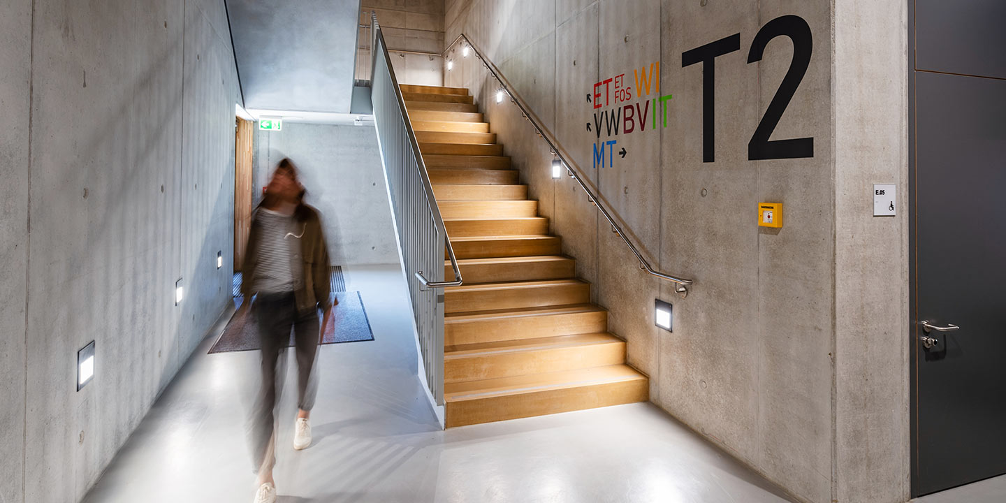 Innenansicht eines modernen Bürogebäudes mit einer gehenden Person, Treppe, wandmontierten Leuchten und Notausgangsschild