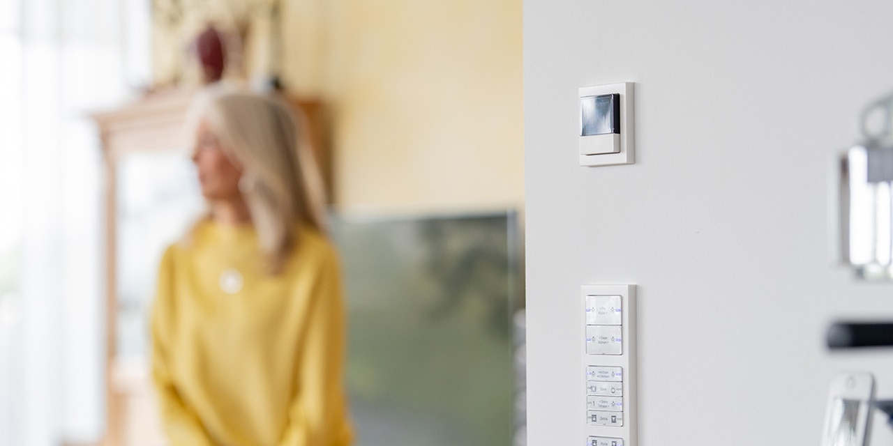 Modernes Zuhause mit verschwommener Person im Hintergrund und Fokus auf wandmontiertem Smart-Thermostat und multifunktionalem Lichtschalterpanel