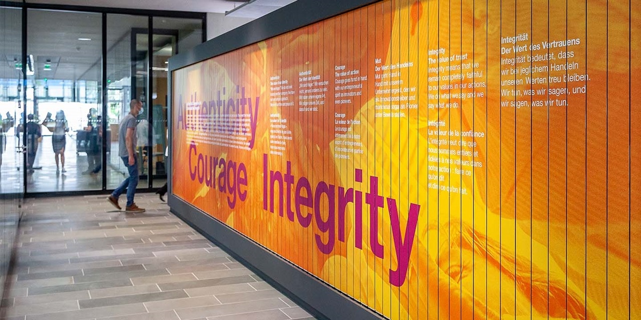 Bürokorridor mit vorbeigehenden Personen an einer inspirierenden Wandgrafik mit den Worten 'Authenticity, Courage, Integrity' auf Englisch, keine sichtbaren elektrischen Geräte