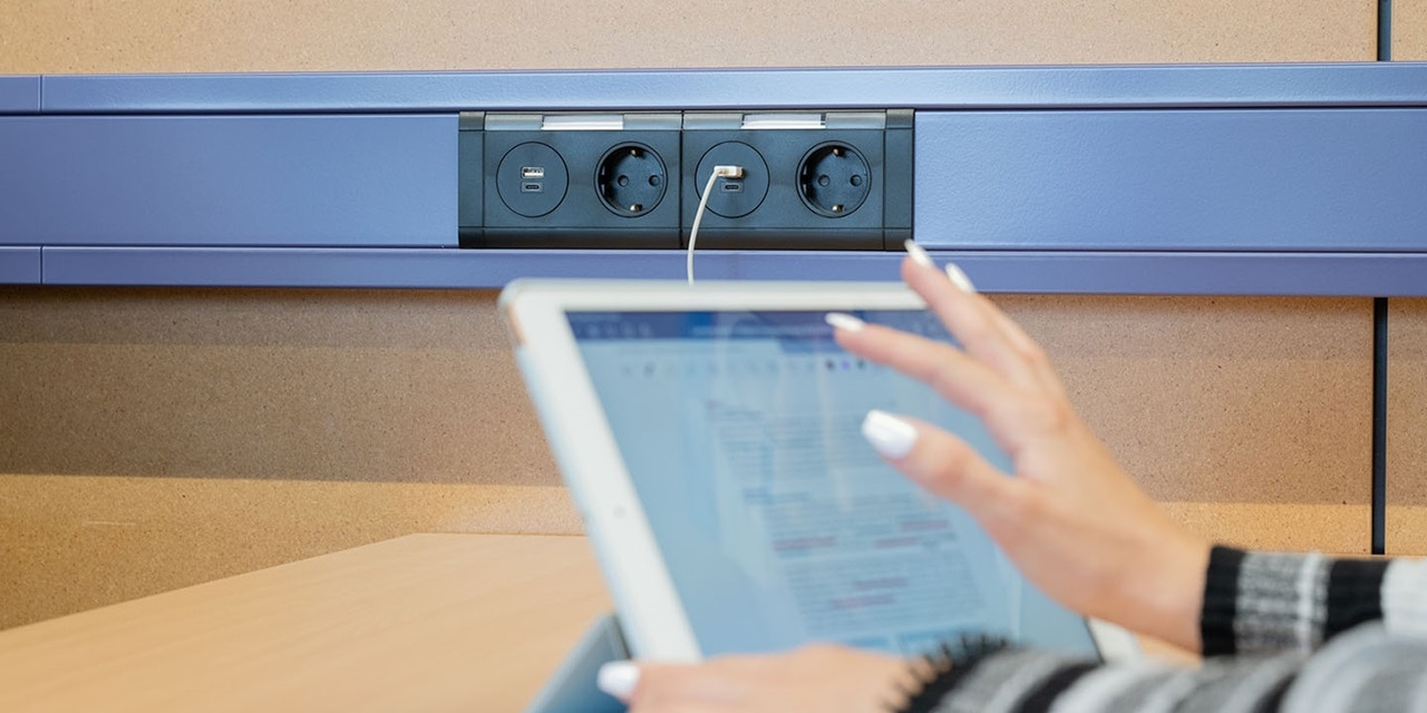 Person benutzt ein Tablet vor einer Steckerleiste mit europäischen Steckdosen auf einer blauen Schiene unter einem Tisch