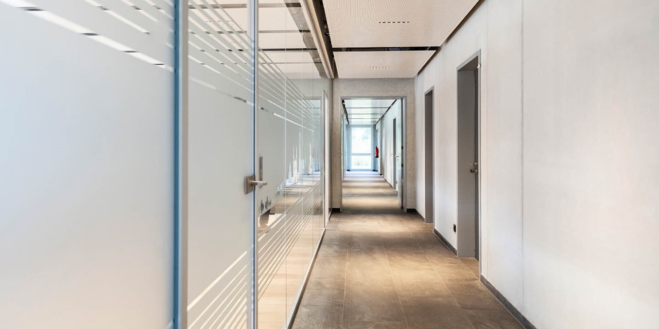 Moderner Büroraum mit Glaswänden und Türen, Einbaubeleuchtung und einer Person am Ende des Flurs