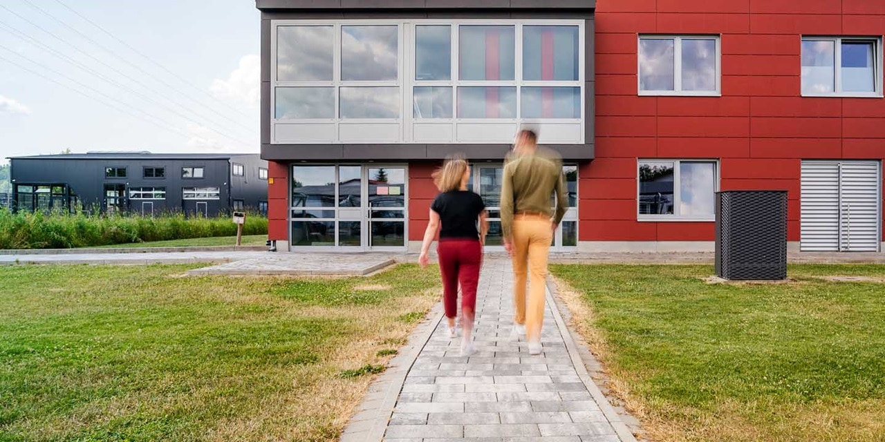 Zwei Personen gehen auf ein modernes Geschäftsgebäude mit roter und grauer Fassade und großen Fenstern zu