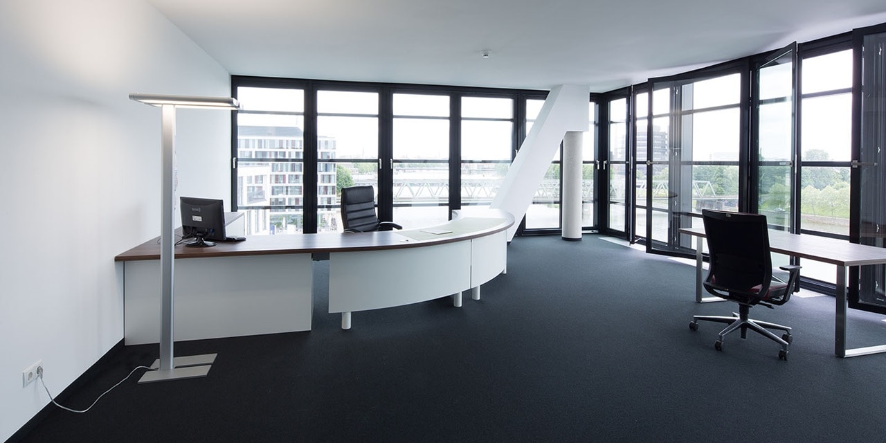 Modernes Büro-Interieur mit Schreibtischen, Bürostühlen und Stehleuchten, große Fenster sorgen für reichlich Tageslicht