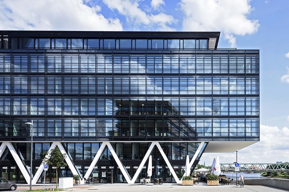 Modernes Bürogebäude mit Glasfassade und markanten weißen Stützen, neben einem Fluss unter klarem Himmel