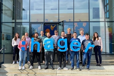 Gruppe von Personen hält Buchstaben, die 'HAGER' vor einem Firmengebäude buchstabieren