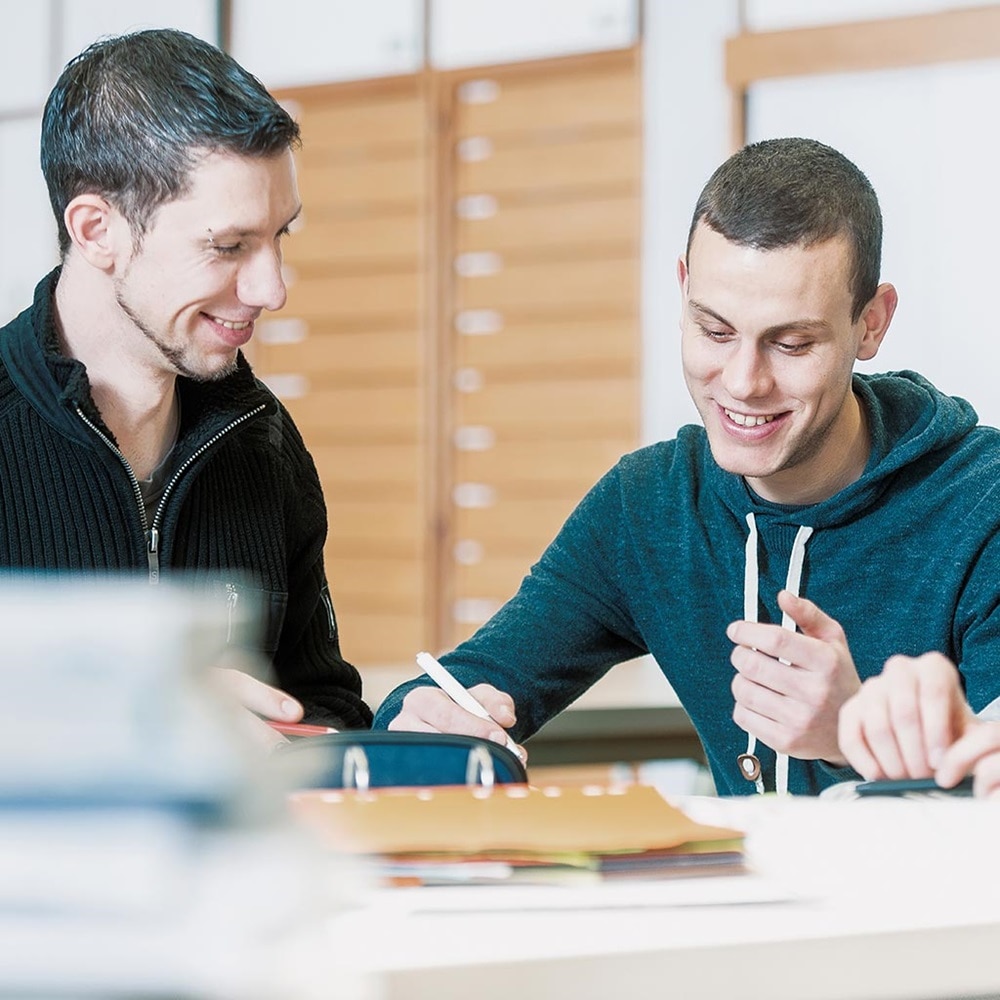 Zwei lächelnde Auszubildende an einem Tisch im Schulsaal, die zusammen lernen, ein Sinnbild für Hager Weiterbilung.